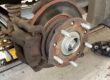 Wheel removed showing a worn brake disc with heavy corrosion and a pronounced lip