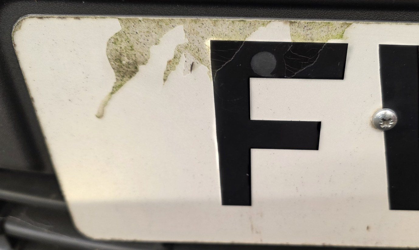 Number plate peeling at the top edge with a fixing screw through a character